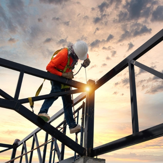 Persona con implementos de seguridad para alturas instalando una estructura de acero para la construcción de techo. De fondo un atardecer.