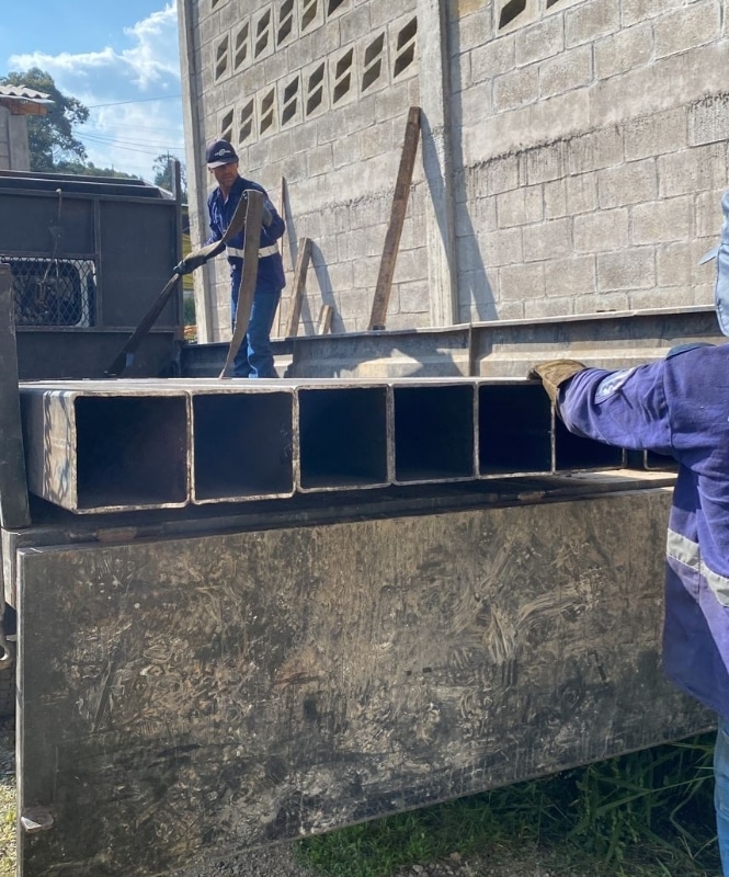 Dos personas amarrando tubos de acero cuadrado en la parte trasera de un camión. La primer persona está de pie encima de la cama del camión tomando las agarraderas, la otra persona está de pie en el piso tocando con una mano uno de los tubos