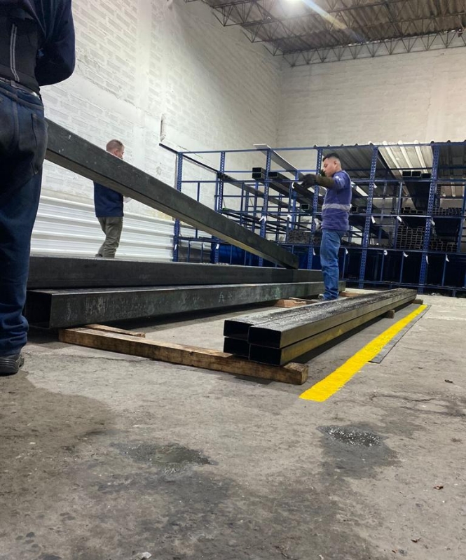 Tres hombres organizando tubería de acero de calidad de forma rectangular en bodega. De fondo estantería con acero.