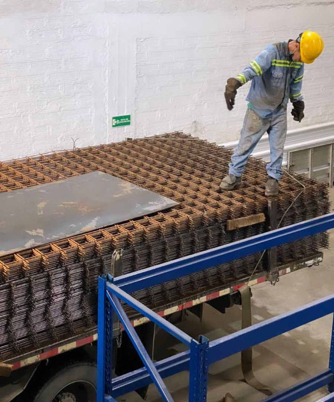 Hombre organizando malla electrosoldada hecha con acero de calidad sobre cama trasera de camión dentro de bodega.